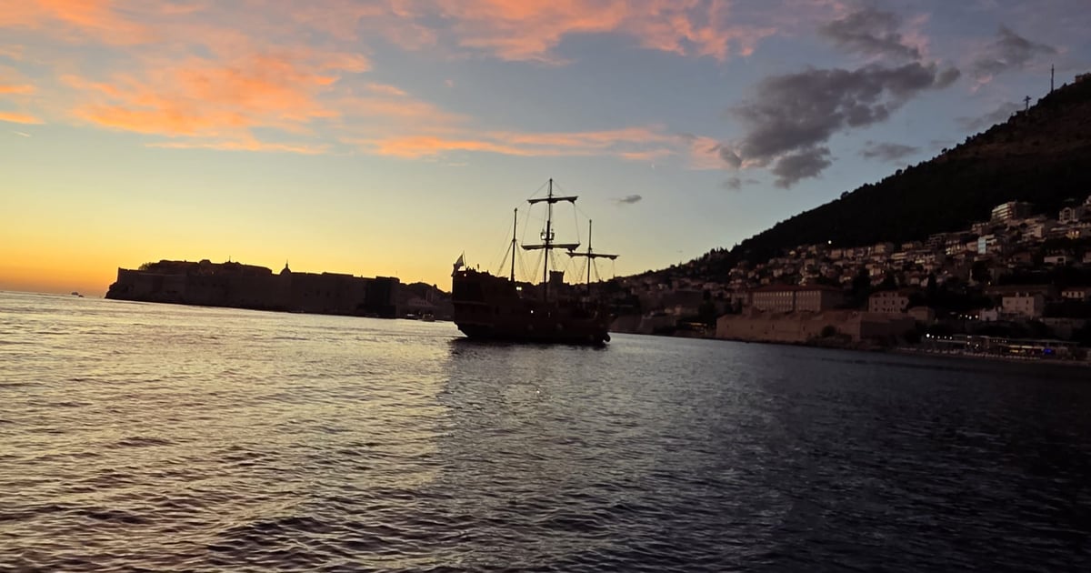 A replica sailing ship in front of Dubrovnik’s walls at dusk, with lights coming on along the hillside