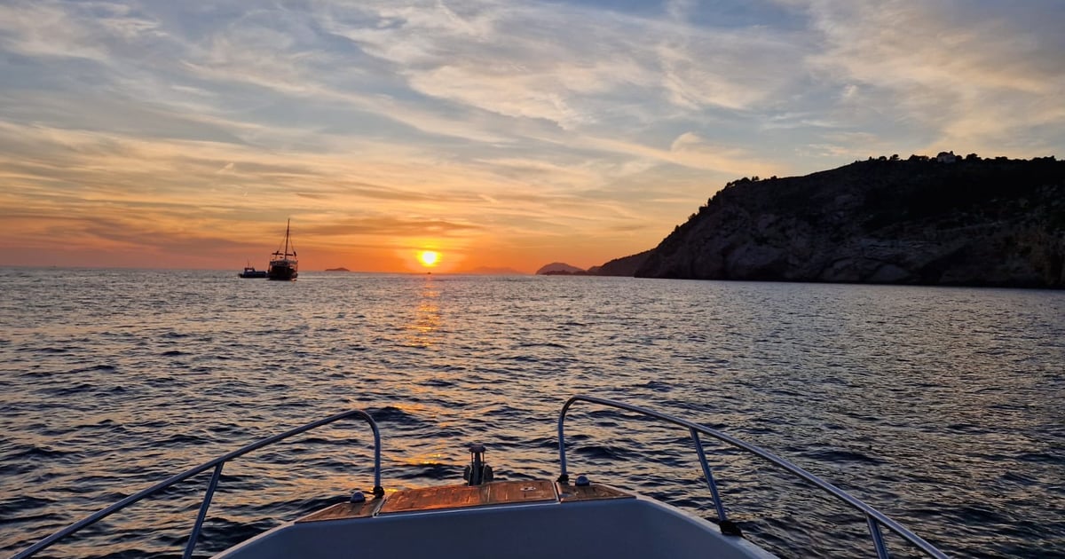 The view over the bow of a small boat: the sun setting into the sea beside a dark headland, with a sailing ship on the horizon