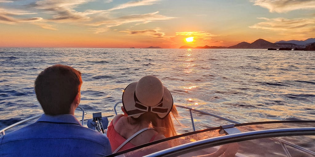 A couple at the bow of a small boat, the woman in a straw sun hat, watching the sunset behind a line of islands off Dubrovnik