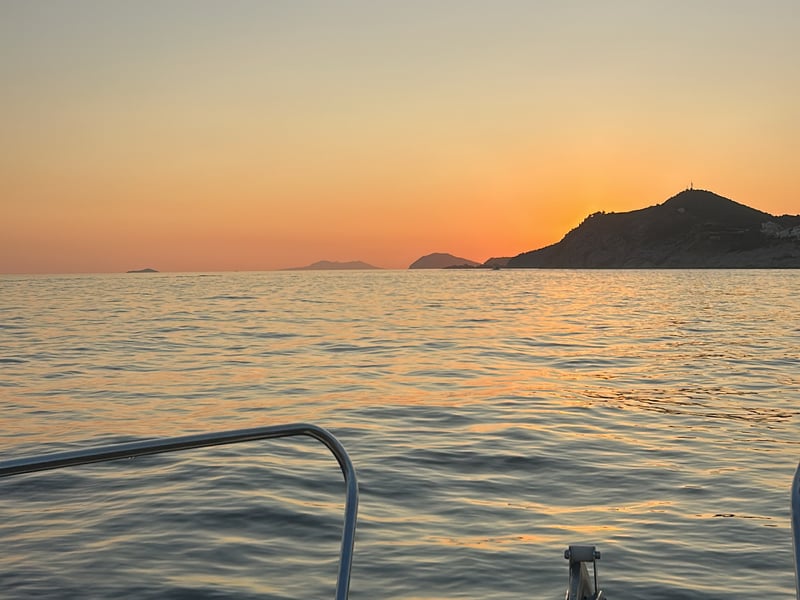 Orange afterglow over the sea and a hilly headland, seen over the bow rail of a small boat