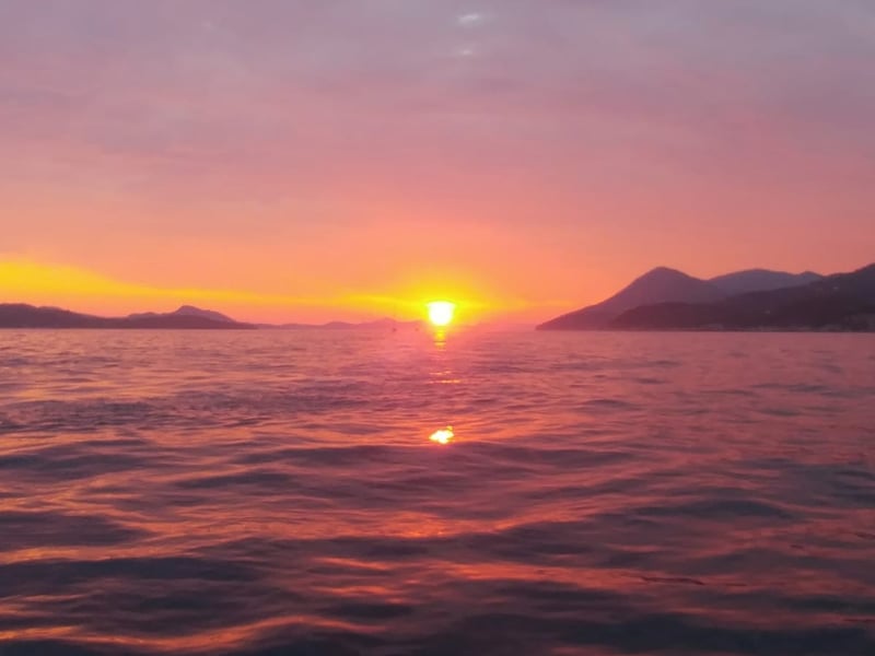 A pink sky over the sea as the sun sets between two lines of islands, seen from the water