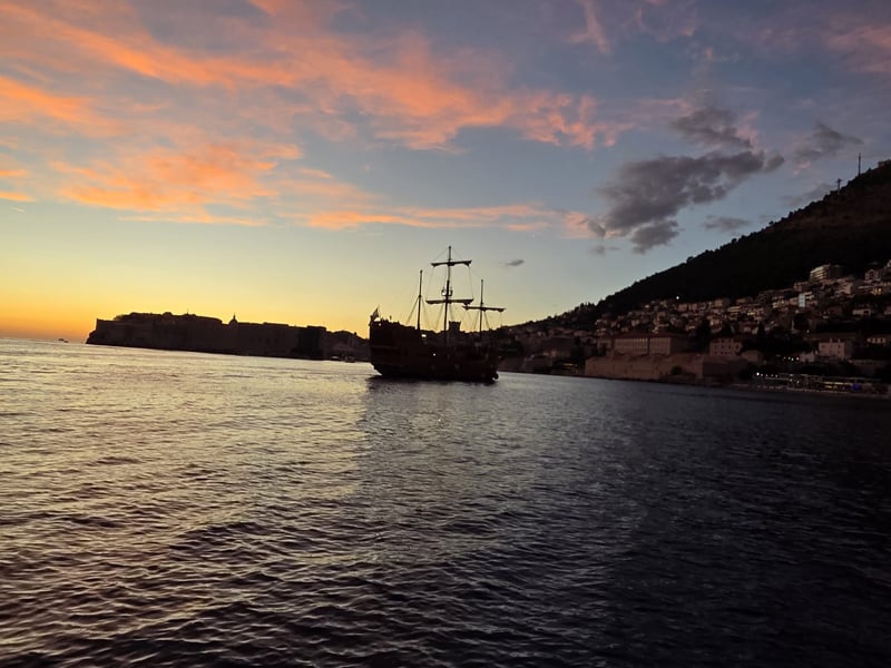 A replica sailing ship in front of Dubrovnik’s walls at dusk, with lights coming on along the hillside