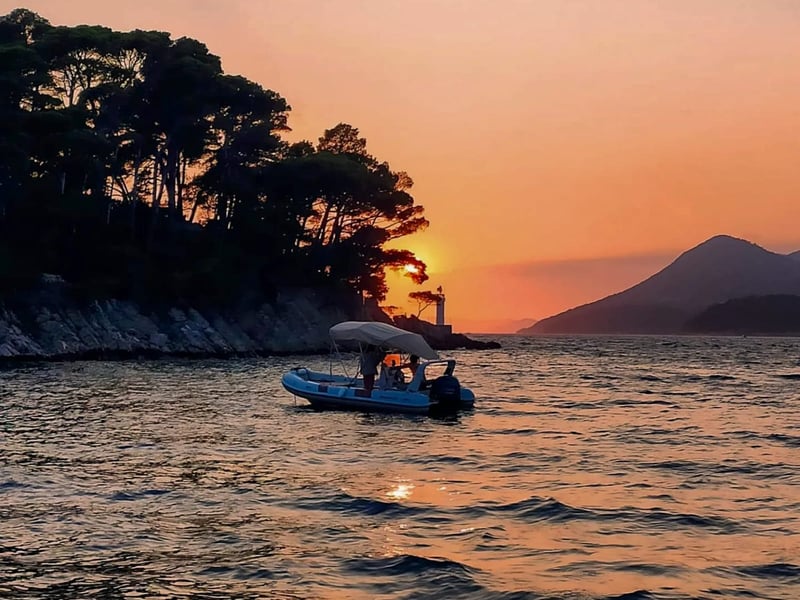 A small boat off a pine-covered island with a little lighthouse, as the sun sets behind the trees