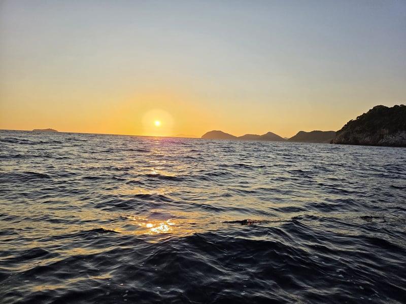 The sun low over the sea, with a line of islands on the horizon and a rocky headland on the right