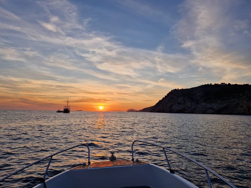 The view over the bow of a small boat: the sun setting into the sea beside a dark headland, with a sailing ship on the horizon